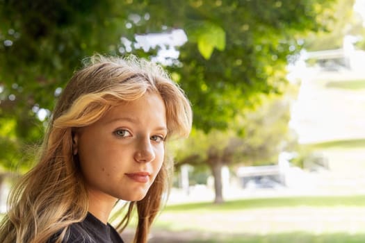 portrait of a teenage girl with blond hair, the girl sits in the park and looks at the camera. High quality photo