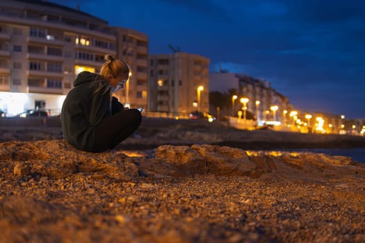 The girl sits on the embankment in the evening and looks into the phone. a girl sits near the sea in the evening close-up, There is a place for an inscription. High quality photo