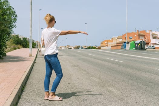 a beautiful girl in a white t-shirt and jeans stands on the road with her back to the camera and stops the car on the road, there is a place for an inscription. High quality photo