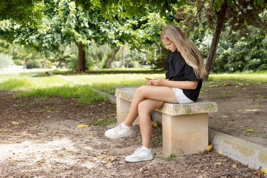 teenage girl in shorts and a black T-shirt sits on a park bench and looks at the phone. High quality photo