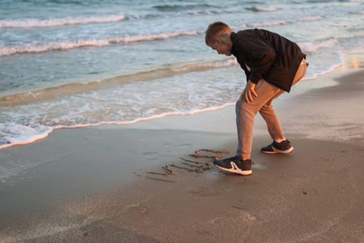 a happy child of European appearance sits on the beach near the sea draws on the sand. A child on vacation sits near the sea. High quality photo