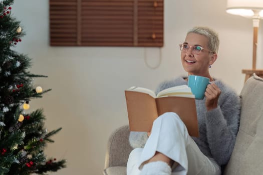 Caucasian senior woman reading book and drinking hot drink and relaxation on couch on Christmas holiday.