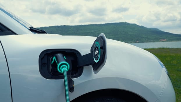 Electric car plug in with charging station, recharge battery by EV charger cable display smart digital battery status hologram with nature background. Futuristic green energy infrastructure.Peruse