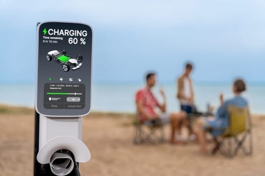 Alternative family vacation trip traveling by the beach with electric car recharging battery from EV charging station with blurred family enjoying the seascape background. Perpetual