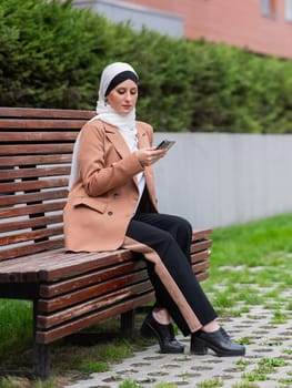 Young woman in hijab uses a smartphone while sitting on a bench outdoors