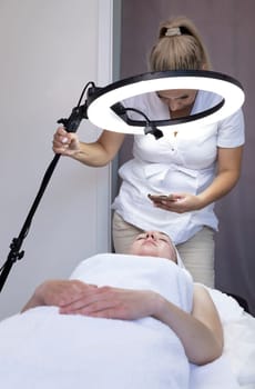Caucasian Woman Cosmetologist Or Beautician Takes Pictures On Phone Of Cosmetology Procedure Result On Young Beautiful Female Patient In Medical Office, Led Selfie Ring Light on Tripod. Vertical