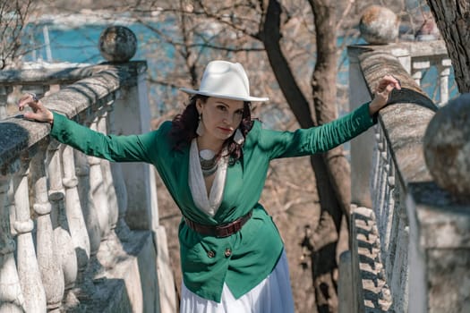 Woman walks around the city, lifestyle. Happy woman in a green jacket, white skirt and hat is sitting on a white fence with balusters overlooking the sea bay and the city