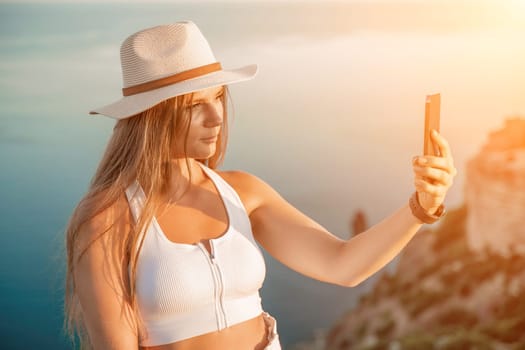 Selfie woman in a hat, white tank top, and shorts captures a selfie shot with her mobile phone against the backdrop of a serene beach and blue sea