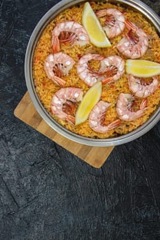 Vertical photo with a top view of a frying pan with paella on a wooden board on a black textured background. flat lay Classic dish of Spain, seafood paella, copy space. soft focus.