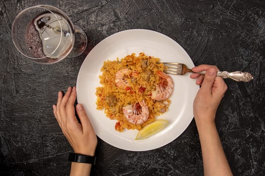 top view of female hands with fork, white plate with paella and shrimp and glass of red wine on black textured background. dinner alone. flat lay with classic dish of Spain, seafood paella,