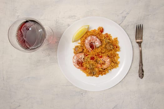 flat lay of a white plate with paella and shrimp and a glass of red wine on a white textured background. Top view classic dish of Spain, seafood paella