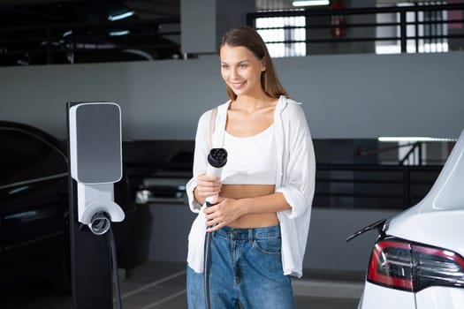 Young woman travel with EV electric car to shopping center parking lot charging in downtown city showing urban sustainability lifestyle by green clean rechargeable energy of electric vehicle innards