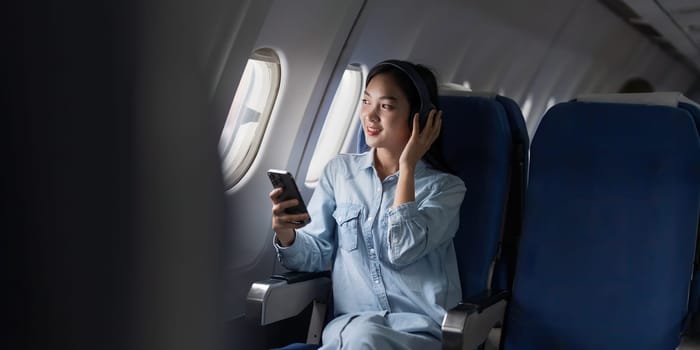 Young female listening favourite song during flight in first class cabin using smartphone, woman entertain on airplane board enjoy music in headphone from smartphone.