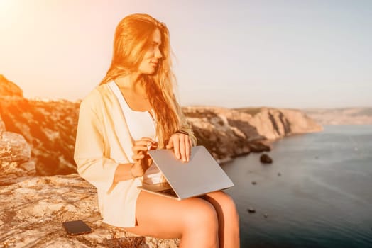 Successful business woman in yellow hat working on laptop by the sea. Pretty lady typing on computer at summer day outdoors. Freelance, travel and holidays concept.