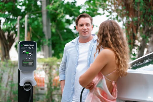 Happy and lovely couple with eco-friendly conscious recharging electric vehicle from EV charging station. EV car technology utilized as alternative transportation for future sustainability. Synchronos