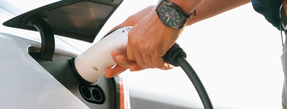 Closeup hand put EV charger to recharge electric car's battery in city commercial parking lot. Rechargeable EV car for sustainable environmental friendly urban travel. Panorama Expedient