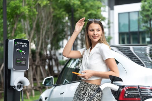 Young woman travel with EV electric car charging in green sustainable city outdoor garden in summer. Urban sustainability lifestyle by green clean rechargeable energy of electric BEV vehicle innards