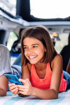 girl smiling happy using mobile phone lying in a camper van, concept of technology and active tourism in nature with children