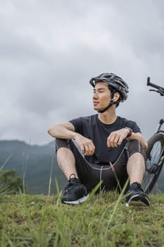 handsome Asian man in sportswear and a bike helmet enjoys listening to music through his earbuds while riding a bike on the country roads on the weekend