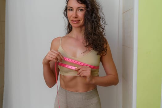 Cropped view of slim woman measuring breasts with tape measure at home, close up. Unrecognizable European woman checks the result of a weight loss diet or liposuction indoors. Healthy lifestyle..