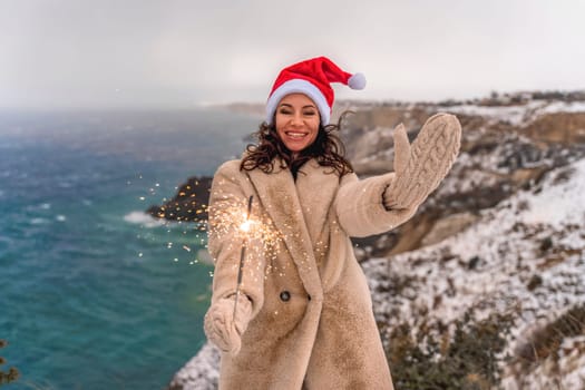 Outdoor winter portrait of elegant happy smiling woman in santa hat, light faux fur coat holding sparkler, posing against sea and snow background