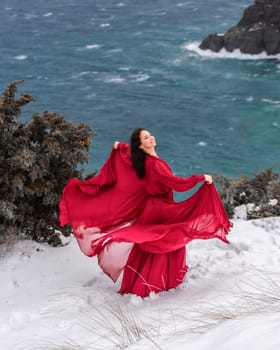 Woman red dress snow sea. Happy woman in a red dress in the snowy mountains by the emerald sea. The wind blows her clothes, posing against sea and snow background