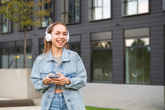 A young woman in headphones and smiling her gaze towards the camera, holding a phone in hand.