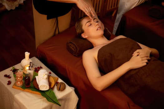 Caucasian woman enjoying relaxing anti-stress head massage and pampering facial beauty skin recreation leisure in warm candle lighting ambient salon spa in luxury resort or hotel. Quiescent