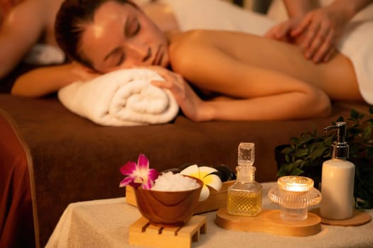 Caucasian couple customer enjoying relaxing anti-stress spa massage and pampering with beauty skin recreation leisure in warm candle lighting ambient salon spa at luxury resort or hotel. Quiescent