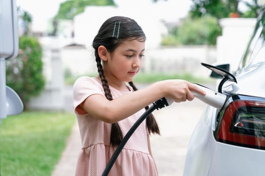 Happy little young girl learn about eco-friendly and energy sustainability as she recharge electric vehicle from home EV charging station. EV car and sustainable future generation concept. Synchronos