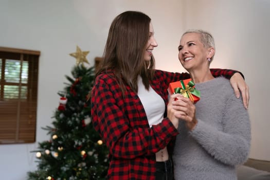 Daughter hug elderly mother with gift box at home in front of tree decorated with Christmas decorations.