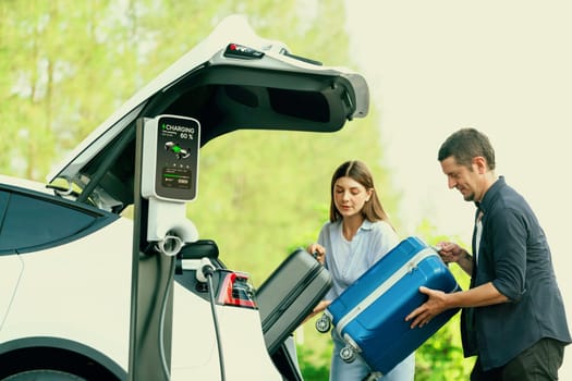 Lovely young couple recharging battery for electric car while relaxing and sitting on the trunk during road trip travel EV car in autumnal forest. Eco friendly travel on vacation during autumn. Exalt