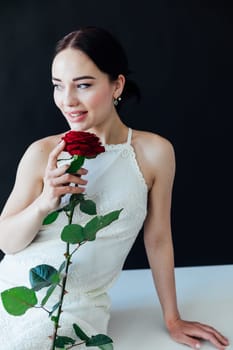 beautiful brunette in white dress with a red rose sits on a table against a black wall