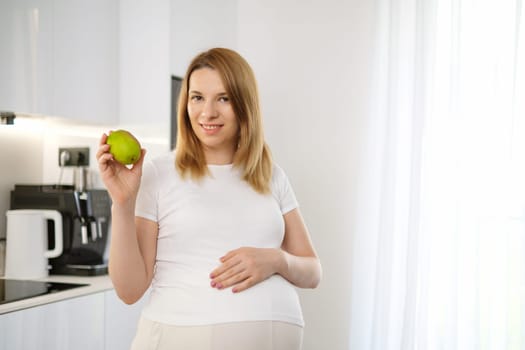 Beautiful blonde pregnant woman holding a pear and tough her belly standing in the kitchen.