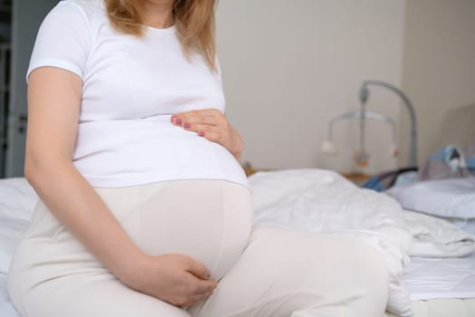 Pregnant woman sits on the bed with a baby crib adorned with hanging toys on the background.