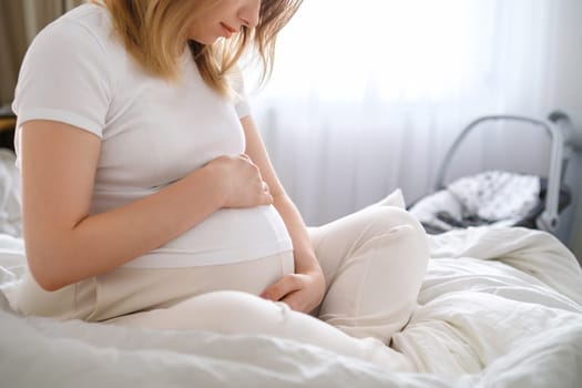 Pregnant woman in light clothes sits on the bed and gently hugs her belly.