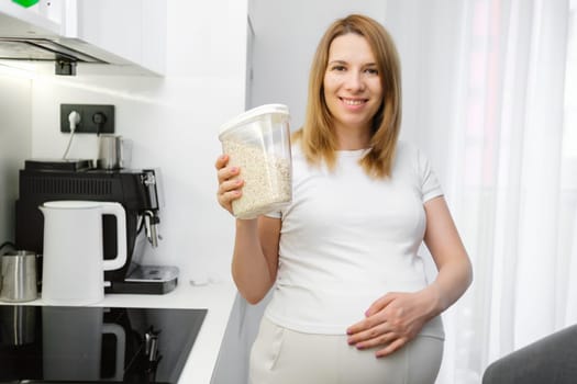 Beautiful pregnant woman holding a container with oats in the kitchen. Diet and vitamins during pregnancy.