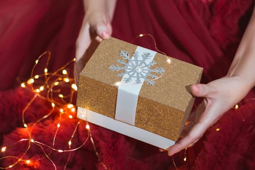 A gift in women's hands on the background of a red dress with lights from a garland