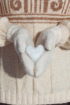 A girl in a beige sweater holds in her hands, dressed in white mittens, a heart made of snow close-up. Postcard for Valentine's Day