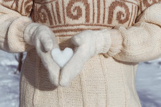 A girl in a beige sweater holds a heart made of snow in her hands, dressed in white mittens, from the side. A gift for Valentine's Day. Postcard with copy space