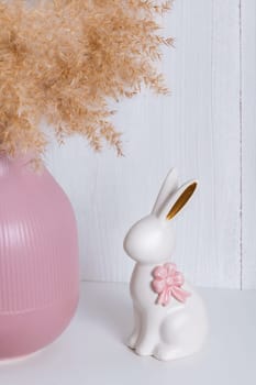 A white porcelain rabbit with a pink bow on a shelf next to a pink vase and a reed