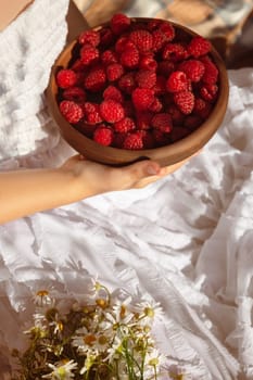 A girl in a white dress holds a clay plate with red ripe raspberries, next to a bouquet of daisies