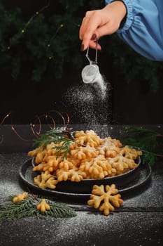 A woman's hand sprinkles powdered sugar on a sweet treat brushwood in the form of snowflakes