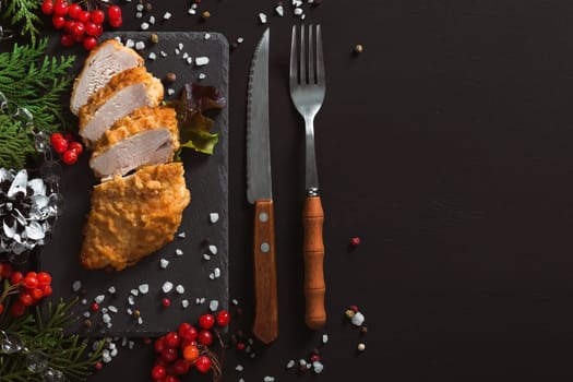 White chicken meat fried in batter on a black background with Christmas decorations and cutlery. The chicken breast is cut into pieces. Top view, copy space