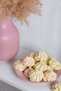 Yellow airy meringue on a pink plate near a vase with reeds on a white background
