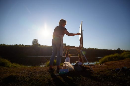 Adult female artist painting picture near water of river or lake in nature and girls in white sundress and flower wreath. Artist and models posing in holiday of Ivan Kupala in nature at sunset