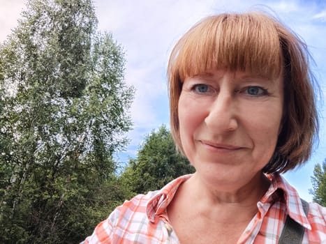 Funny adult girl posing in nature and blue sky on background. A middle-aged woman takes a selfie in the landscape. City tourist on vacation on picnic