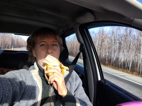 Portrait of female driver in solo journey. Adult mature middle aged woman holding steering wheel and a banana. Eating while driving for to stay awake. Lady girl with food while travel or trip by car