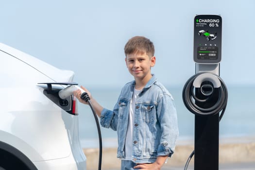 Little boy recharging eco-friendly electric car from EV charging station. EV car road trip travel by the seashore by alternative vehicle powered by clean renewable and sustainable energy. Perpetual