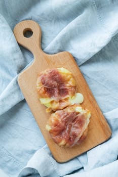Two pieces of camembert cheese wrapped in prosciutto baked in the oven, on a wooden cutting board on a light blue fabric background